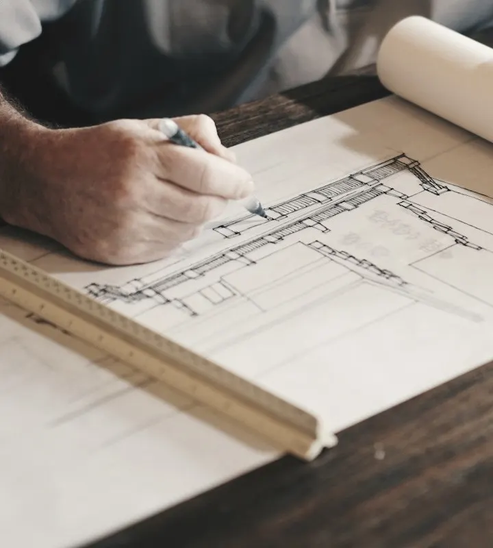 Hands marking a building plan beside a folding ruler on a wooden desk
