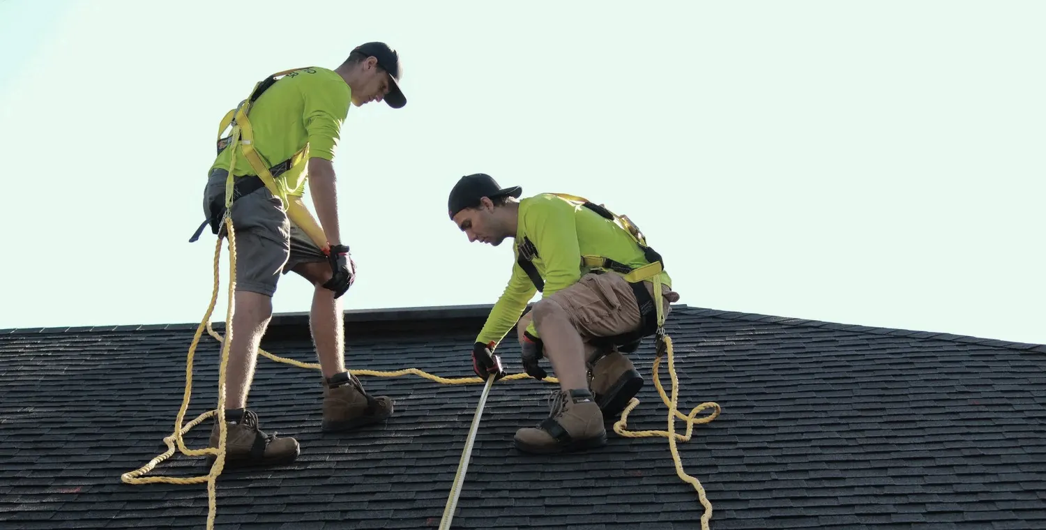 Two workers in safety harnesses measuring a shingle roof